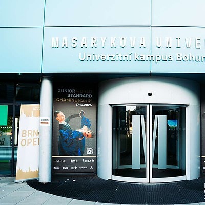 Vstup do univerzitního kampusu Masarykova univerzita v Brně s plakáty o Mistrovství světa juniorů 2026 ve standardu WDSF.