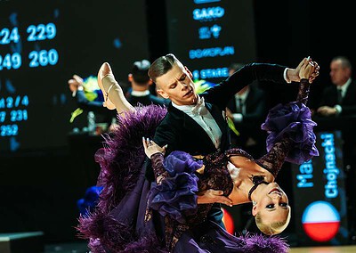 Dva tanečníci v elegantním tanci na tanečním parketu během Mistrovství světa v Brnu, s velkým displejem v pozadí ukazujícím výsledky a statistiky soutěže.