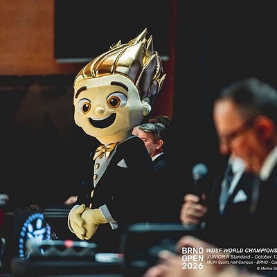 Maskot BrnoOpen 2026, postava s výrazným zlatým účesem a úsměvem, na sportovní akci v Brně, s dalšími účastníky v pozadí.