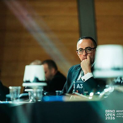 Muž v obleku a brýlích sedí u stolu na badmintonové akci BrnoOpen 2026, s dalšími lidmi v pozadí, v moderním prostředí s dřevěnými stěnami.
